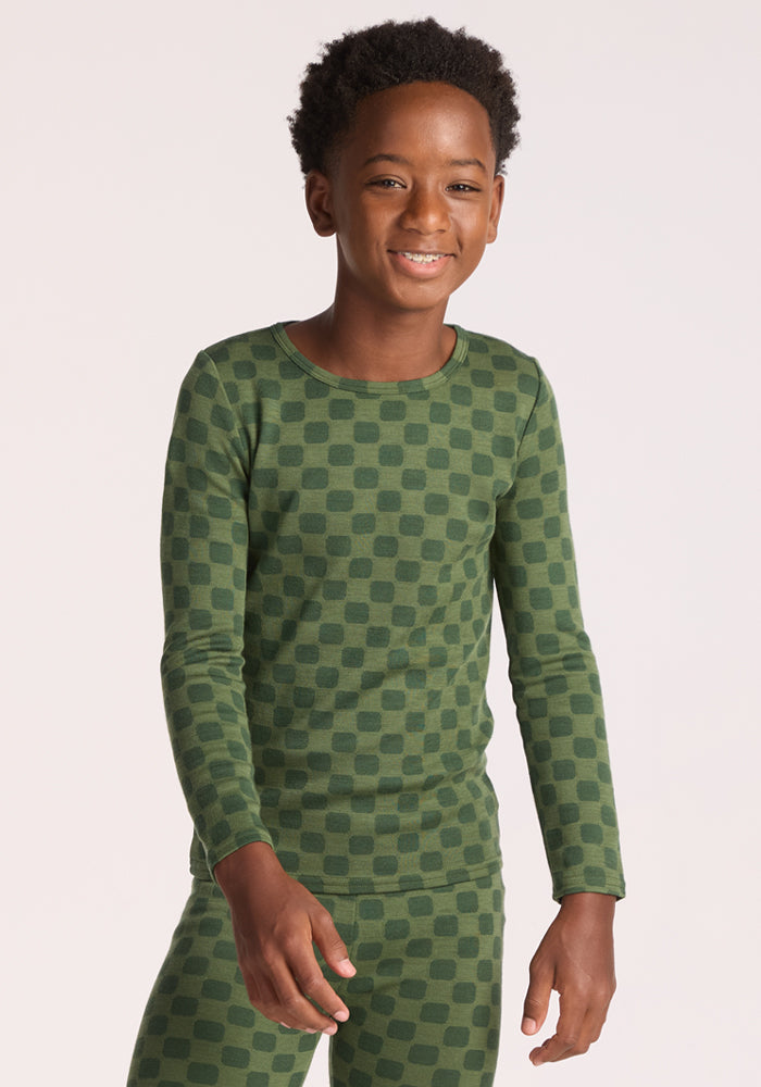 A smiling child wearing the Woolx Kids Explorer Base Layer Top in Frog Hop stands against a plain light background. | Banyan is wearing a size LRG.