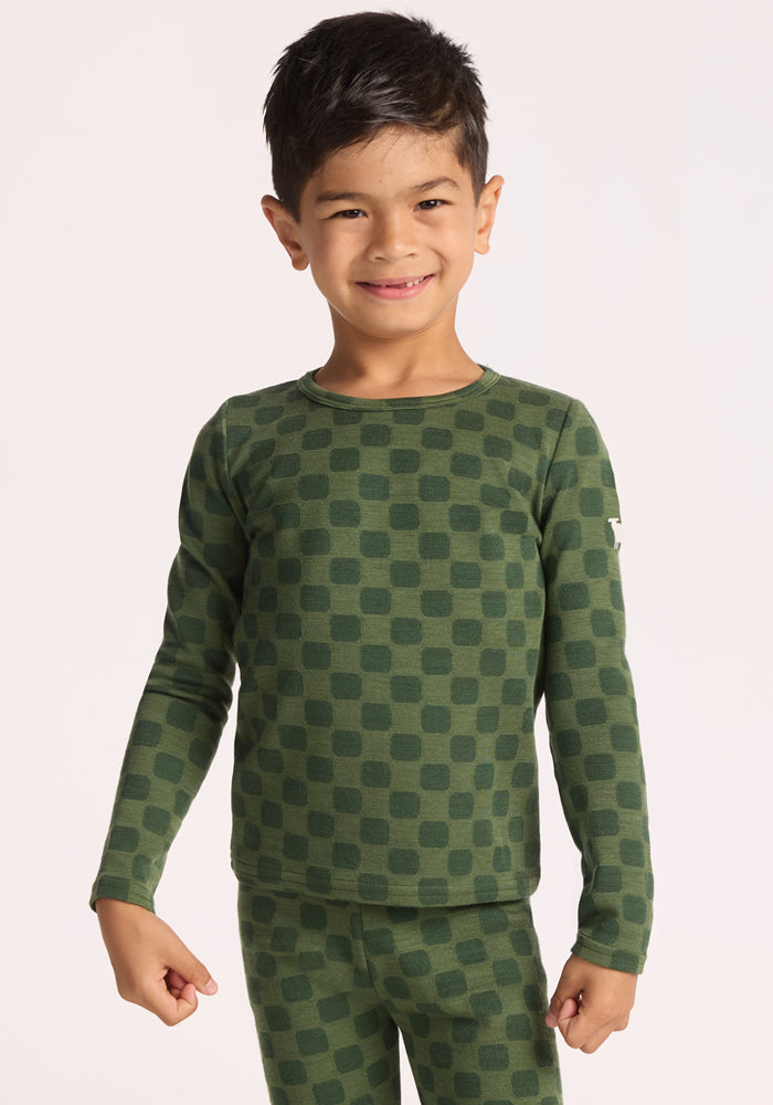 A smiling young child with dark hair wears the Woolx Kids Explorer Base Layer Top - Frog Hop, a cozy and breathable Merino wool thermal featuring a green checkered pattern, standing against a plain light background. | Leo is wearing a size XSM.