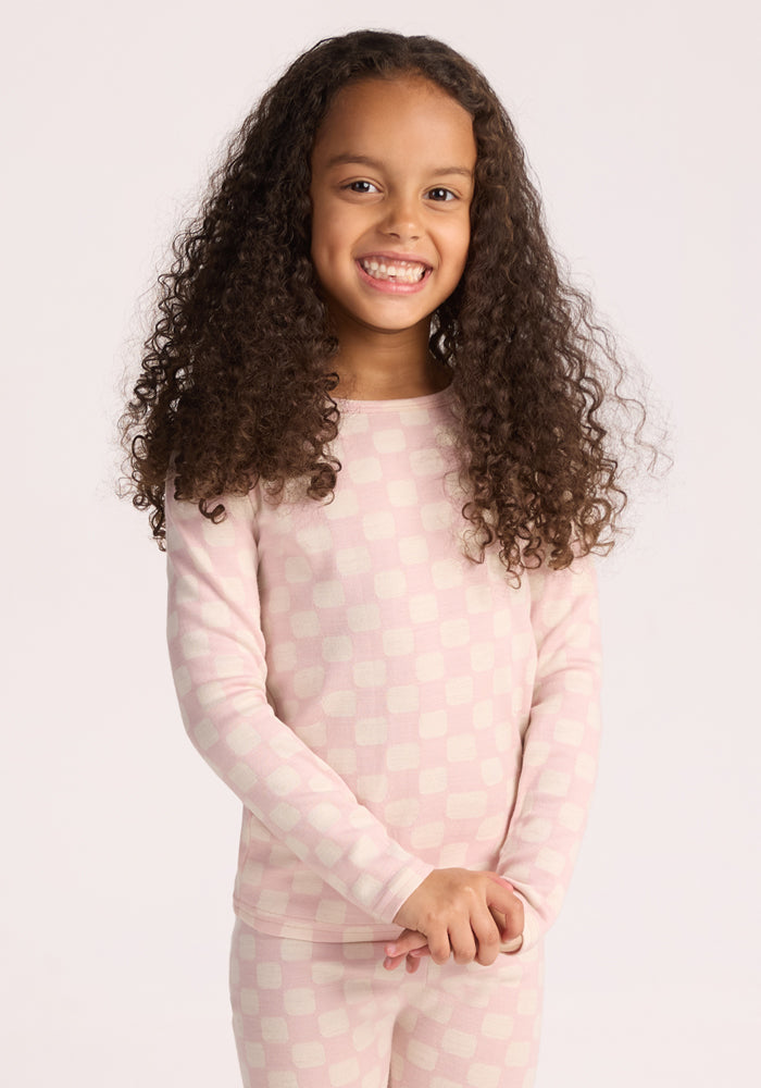 A young girl with long curly hair smiles against a plain background, wearing the Woolx Kids Explorer Base Layer Top in Hopscotch Pink—long-sleeved, cozy, and moisture-wicking—with matching checkered pants for warmth and comfort. | Penny is wearing a size SML.