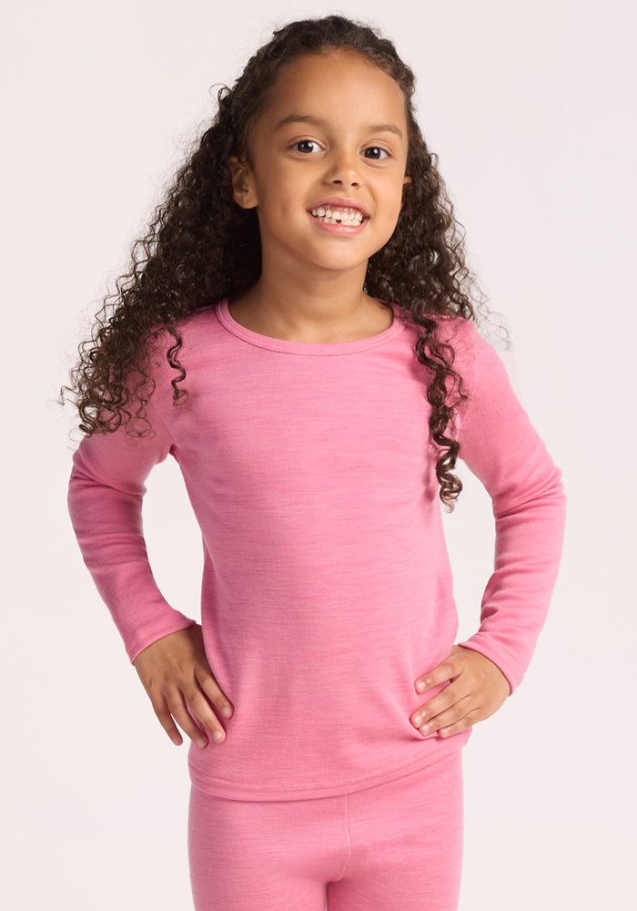 A young child with long curly hair smiles, wearing the Woolx Kids Explorer Base Layer Top in Pinky Promise, standing with hands on hips against a plain light background. | Penny is wearing a size SML.