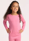 A young child with long curly hair smiles, wearing the Woolx Kids Explorer Base Layer Top in Pinky Promise, standing with hands on hips against a plain light background. | Penny is wearing a size SML.