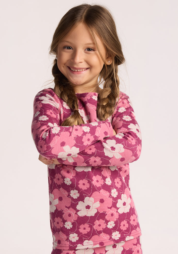 A young girl with long brown hair in braids smiles with arms crossed, wearing the Woolx Kids Explorer Base Layer Top - Rosie Posie, inspired by Merino wool base layers, against a plain light background. | Aria is wearing a size XSM.