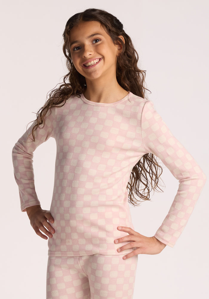 A young girl with long curly hair smiles while posing in the Woolx Kids Explorer Base Layer Top in Hopscotch Pink, featuring long sleeves and matching pants, against a plain light background. | Hanna is wearing a size LRG.