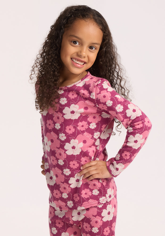 A young girl with long curly hair stands smiling, hands on hips, wearing the Woolx Kids Explorer Base Layer Top in Rosie Posie. She pairs it with matching pants, featuring a pink and white floral pattern against a plain light background. | Penny is wearing a size SML.