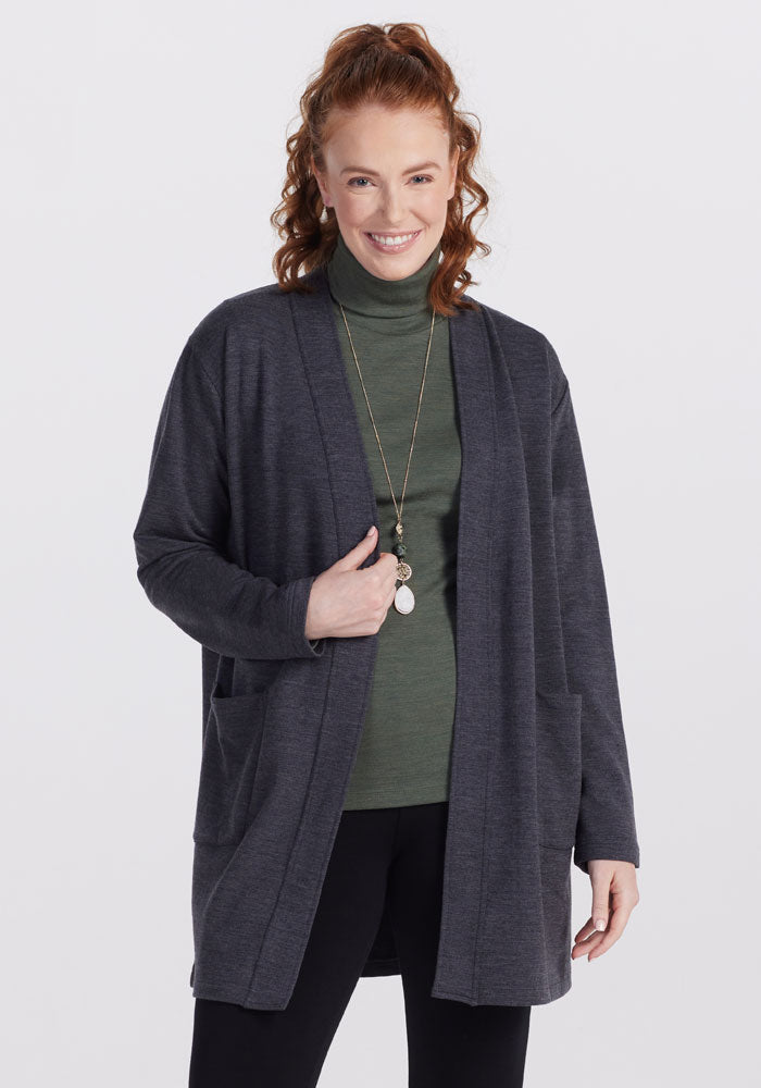 A smiling person with curly red hair wears the Woolx Kinsley Cardigan in Pebble Grey Melange, a green turtleneck, and black pants. They accessorize with a long necklace and have one hand in a cozy pocket, standing against a plain white background.