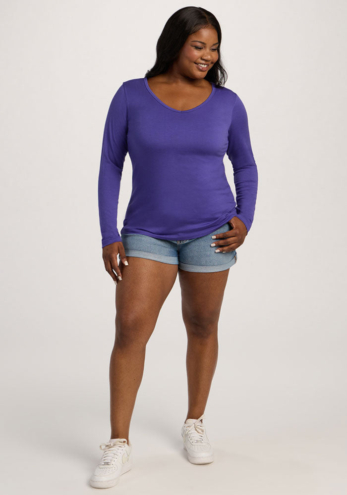 Woman wearing the Woolx Layla V Neck Top in Berry Indigo, denim shorts, and white sneakers stands smiling.