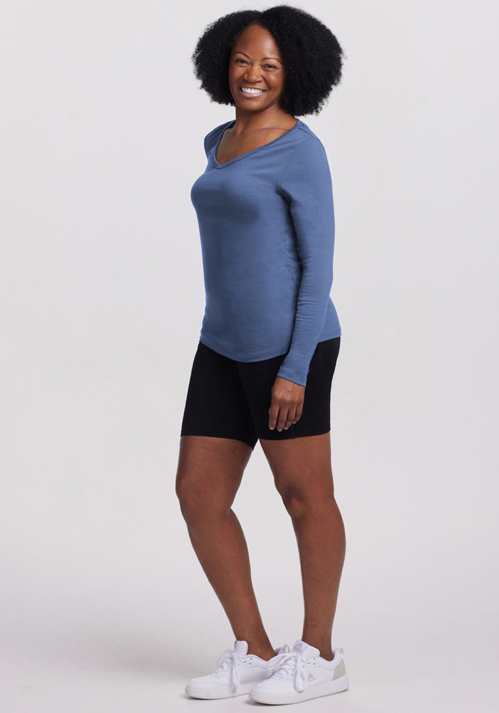 Smiling woman wears the Woolx Layla V Neck Top in Skyline, paired with black shorts and white sneakers.