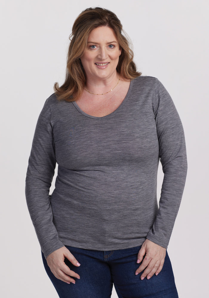 A person with long, light brown hair smiles at the camera, wearing the Woolx Layla V Neck Top in Graphite Heather and blue jeans, standing against a plain white background. | Cambre is 5'11", wearing a size XL