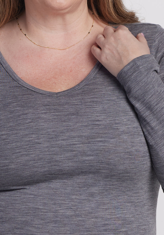 A person is shown from the shoulders down wearing a Layla V Neck Top in Graphite Heather by Woolx. One hand touches their neck, showcasing a delicate gold necklace adorned with small beads. The skin appears slightly flushed.