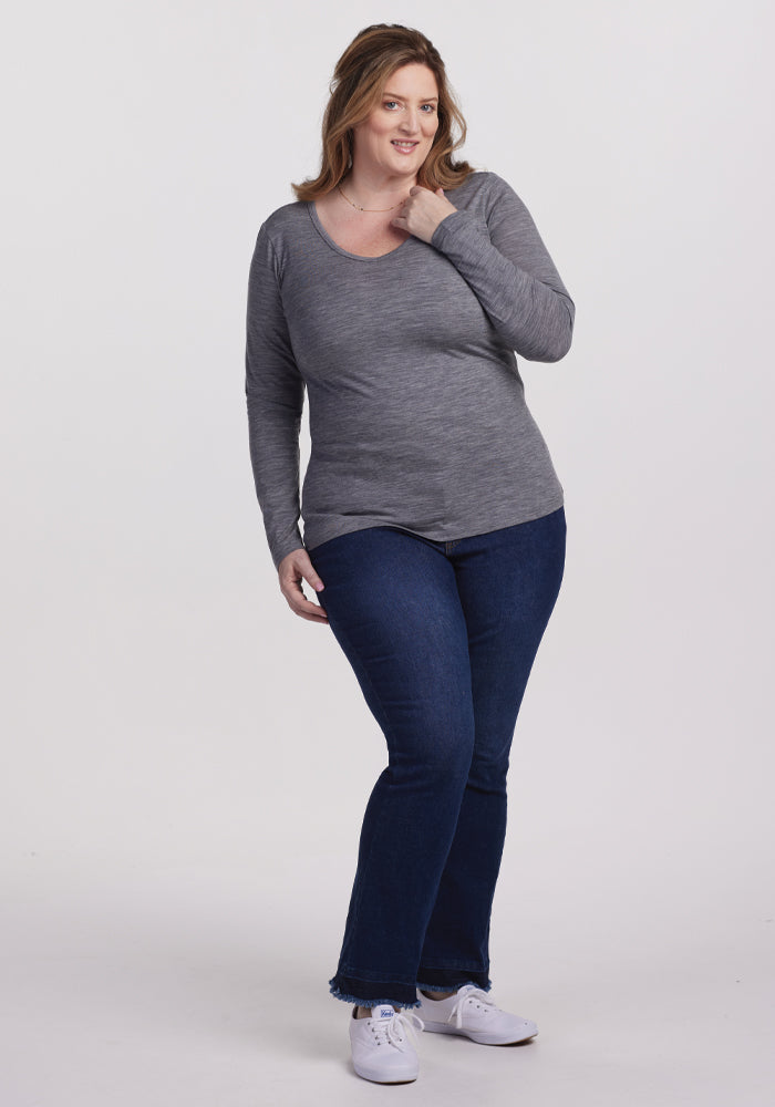 A person with long hair stands against a plain white background, wearing a gray odor-resistant Layla V Neck Top in Graphite Heather by Woolx and dark blue jeans. They rest one hand near their chin and smile, paired with white sneakers.
