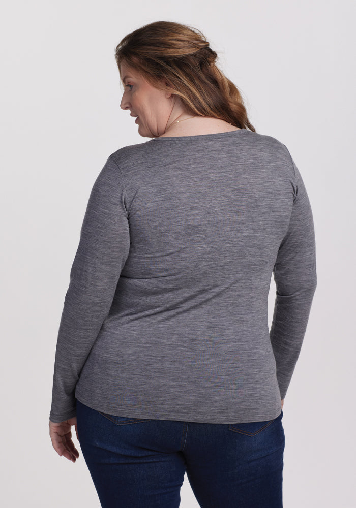 A woman with long brown hair is turned slightly to her left, wearing the Woolx Layla V Neck Top in Graphite Heather and blue jeans against a plain white background.