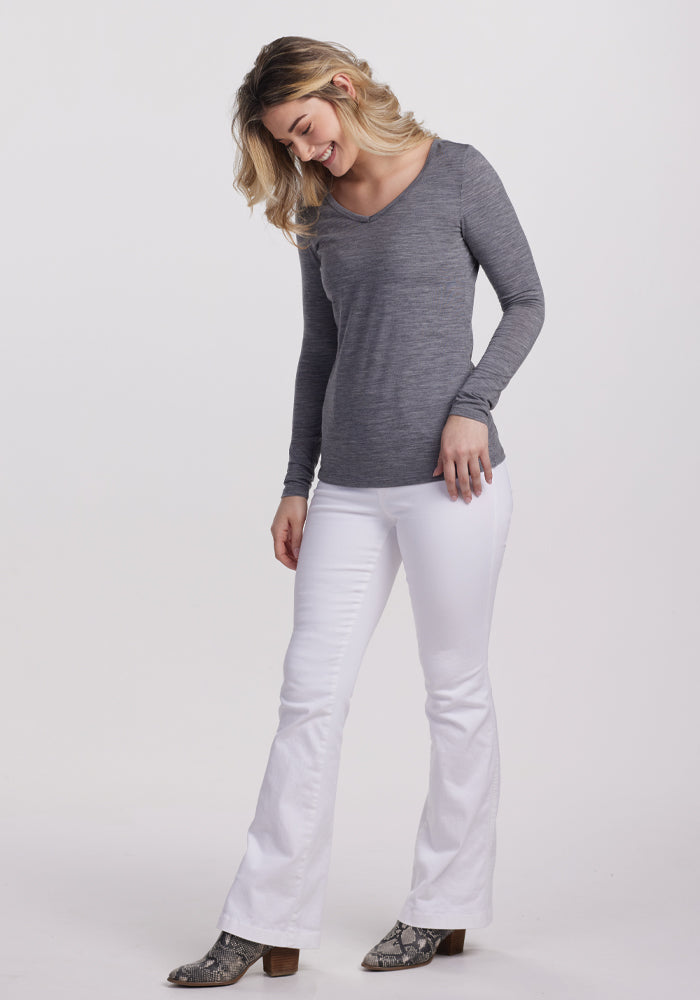 A woman with wavy blonde hair stands on a plain background, wearing the Woolx Layla V Neck Top in Graphite Heather and white bell-bottom pants. She is smiling at the ground, her hands by her sides, accessorized with snakeskin-patterned ankle boots.