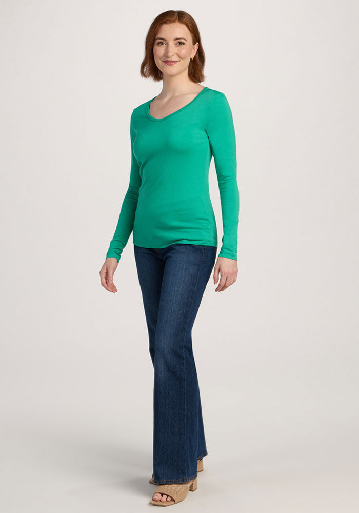 Woman with shoulder-length brown hair wears the Woolx Layla V Neck Top in Jungle, blue jeans, and beige shoes, smiling.