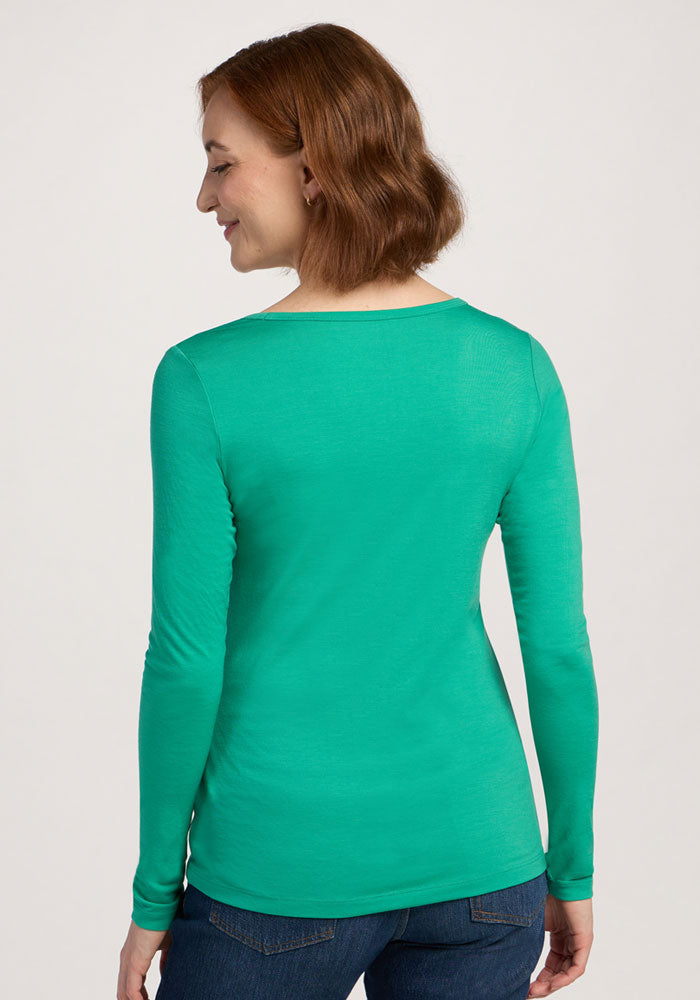 A woman in the Woolx Layla V Neck Top - Jungle and jeans stands facing away, smiling over her shoulder.