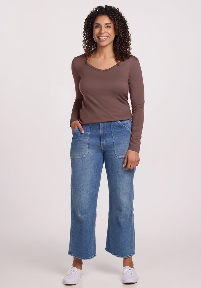 Woman with curly hair in a Woolx Layla V Neck Top in Peppercorn, blue wide-leg jeans, and white sneakers, smiling.