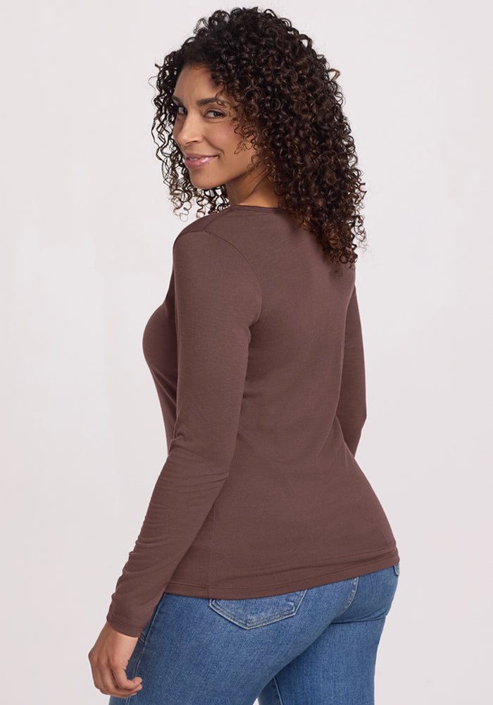 Woman with curly hair in Woolx Layla V Neck Top in Peppercorn and blue jeans, smiling over her shoulder.