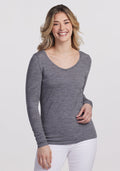 A smiling woman with wavy hair wears the Woolx Layla V Neck Top in Graphite Heather and white pants. She stands against a plain background, resting one hand at her waist and the other by her side, enjoying the odor-resistant comfort of her attire. | Ariana is 5'11", wearing a size S
