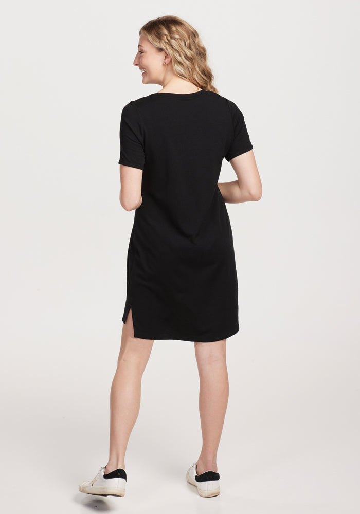 A woman with wavy hair stands with her back to the camera, smiling over her shoulder in a Woolx Lexie Dress & Cover-up - Black and white sneakers against a light gray background, highlighting the dress's versatile elegance.