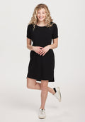 A person with curly hair smiles against a white background, wearing a Woolx Black Lexie Dress & Cover-up paired with white sneakers. Their hands are clasped in front, exuding effortless style and comfort. | Jordan is 5’6”, wearing a size S