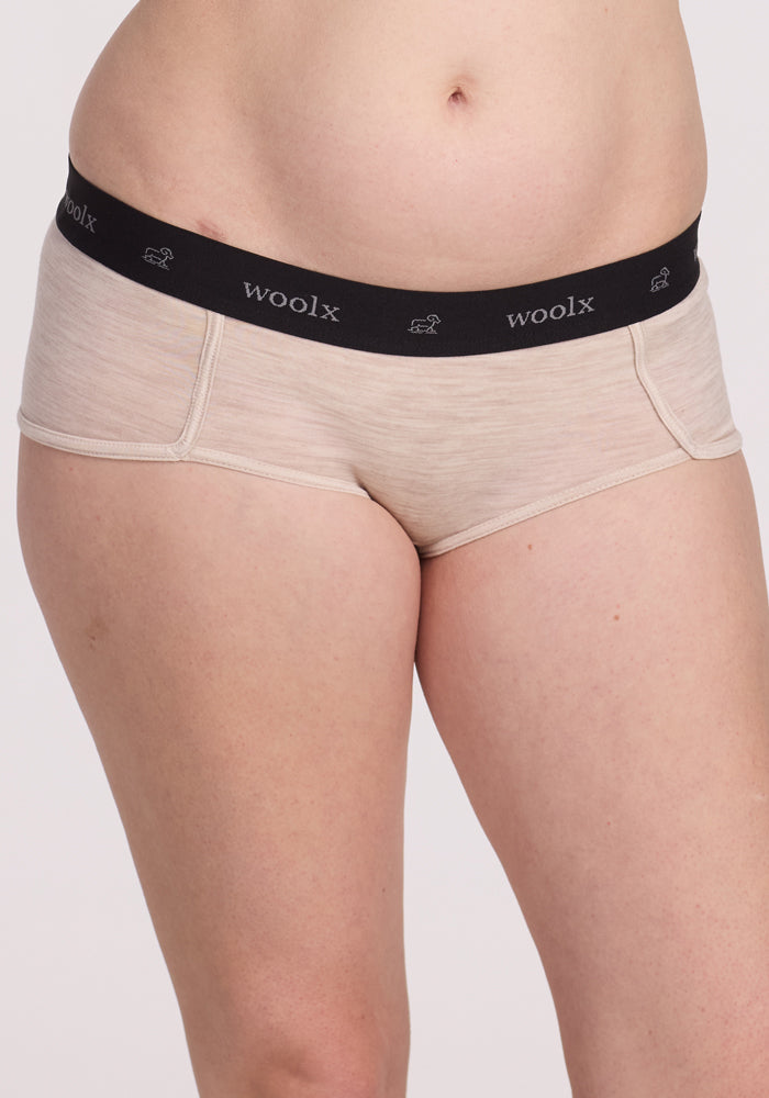 A person wearing Lila Boy Shorts in Cream Heather from Woolx features light beige color and a black waistband with the "Woolx" logo, highlighting sporty construction. The close-up from waist to upper thigh showcases their merino wool blend for all-day comfort against a plain backdrop.