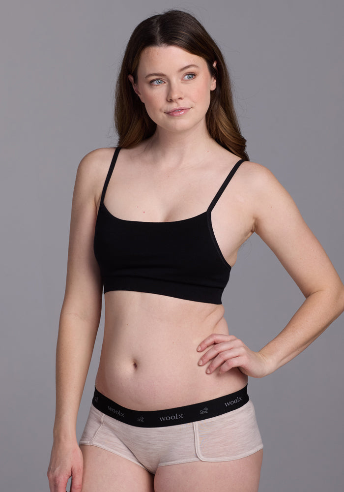 A woman with long brown hair stands against a gray background, wearing a black sleeveless crop top and Woolx's Lila Boy Shorts in Cream Heather. Her sporty look is accentuated by her neutral expression as she gazes slightly to the side.