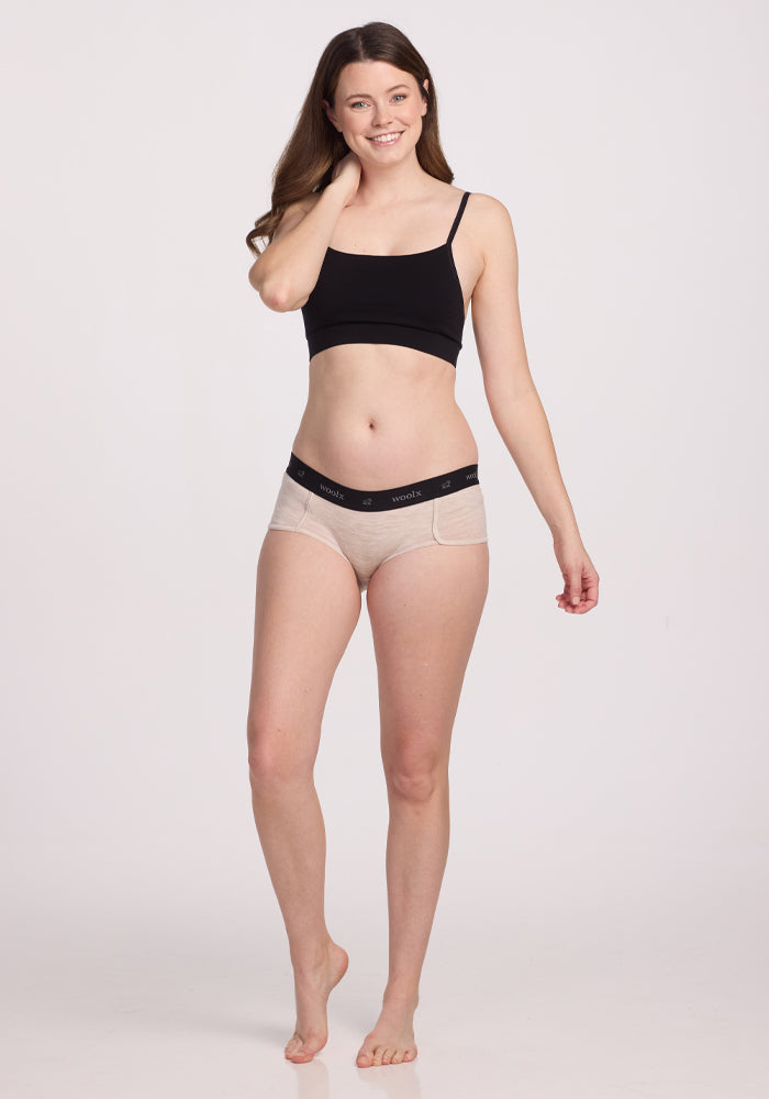 A person with long hair, barefoot and smiling warmly, is wearing a black crop top and the Lila Boy Shorts in Cream Heather by Woolx, crafted from lightweight fabric, posed directly at the camera against a plain light background.