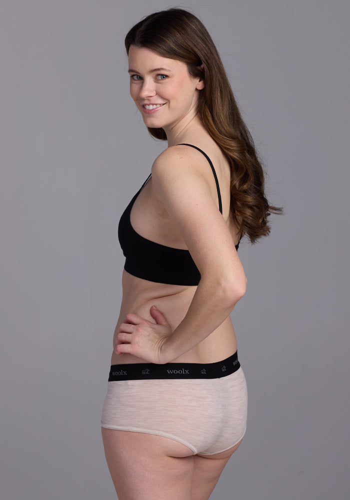 A person with long brown hair is smiling in profile against a gray background, wearing an All-day Comfort black bra and Woolx's Lila Boy Shorts in Cream Heather.