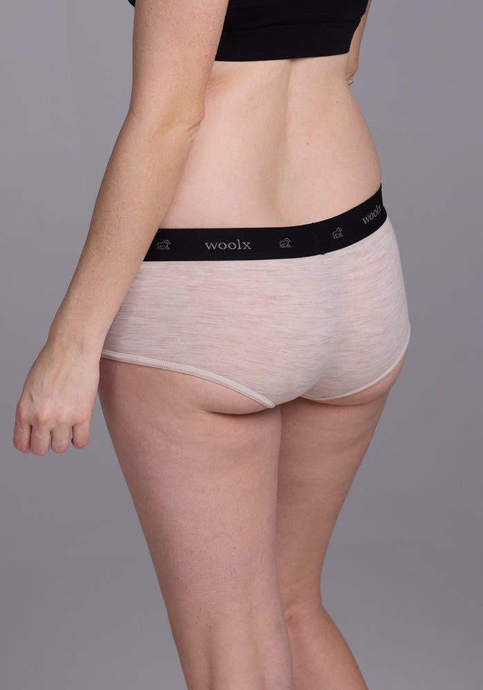 Against a plain gray background, a person models the Lila Boy Shorts in Cream Heather. Made from lightweight Merino wool for comfort, the shorts feature a black waistband that reads "Woolx." The image focuses on their waist and hips from a side view.