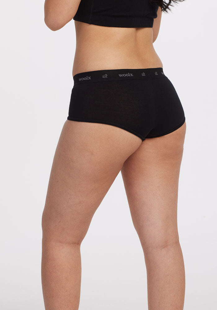 A person is wearing a black top and the Woolx Lila Boy Shorts OLD, crafted from lightweight merino wool, standing against a plain white background. Their back is to the camera, highlighting the fit and style of the clothing.