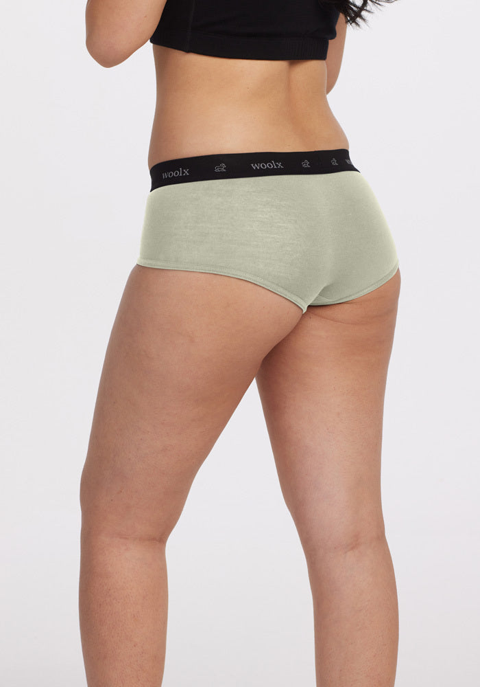 A person is wearing Frosted Mint Lila Boy Shorts with a black waistband featuring "Woolx," and a black crop top. Showcasing the comfort of Woolx DuraLite Fabric, they face away from the camera against a plain white background.