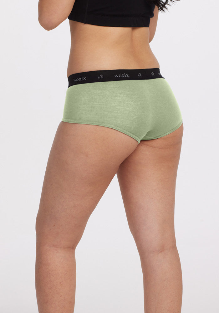 Wearing Woolx's Lila Boy Shorts in Quiet Green, a person stands with their back to the camera, highlighting the comfortable fit certified by Woolx. They pair it with a classic black short-sleeved shirt against a plain white backdrop.