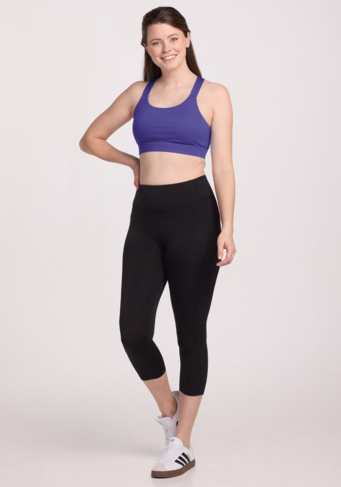 Woman standing, smiling, in a Woolx Livi Bra - Berry Indigo, black leggings, and white sneakers against a plain background.