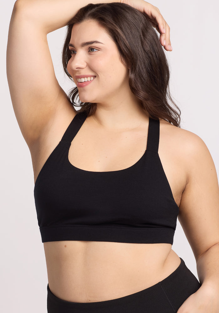 A person with long hair smiles confidently while posing in the Woolx Livi Bra - Black, showcasing its double cross-back straps, paired with leggings. They rest a hand on their head against a plain background, radiating style.