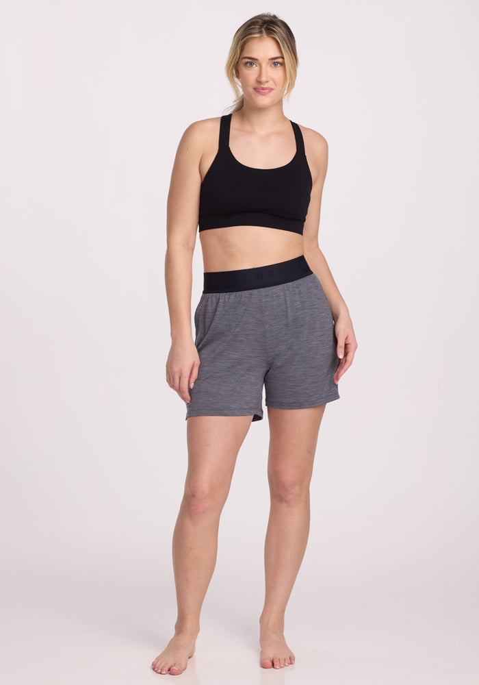 A person stands barefoot in a black Woolx Livi Bra against a white background, wearing gray shorts with a black waistband. Made from an odor-resistant merino wool blend, they have light hair tied back and maintain a neutral expression while looking at the camera.