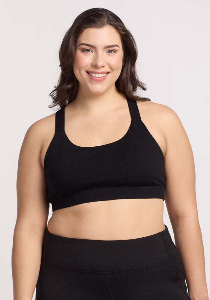 A woman smiles while wearing the Livi Bra - Black by Woolx, featuring double cross-back straps, paired with leggings against a plain white background. | Juliana is 5'10", wearing a size XL