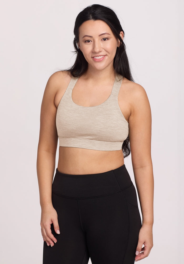 A woman with long dark hair smiles at the camera, wearing the Woolx Livi Bra in Cream Heather and black leggings, standing against a plain light background with her arms relaxed by her sides. | Sarah is 5'8", wearing a size S