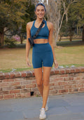 Woman wears Woolx Livi Bra in Majolica Blue, smiling outdoors with a crossbody bag by a brick wall and trees.