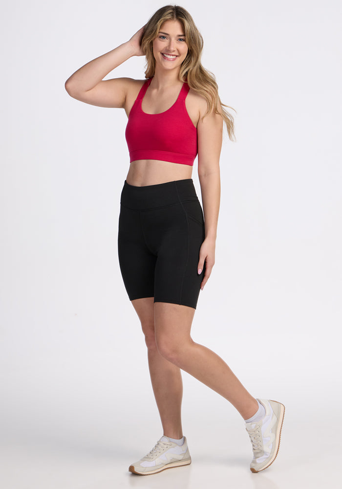 Woman in the Woolx Livi Bra • Final Sale - Poppy Spark, black bike shorts & white sneakers smiles against a white background.