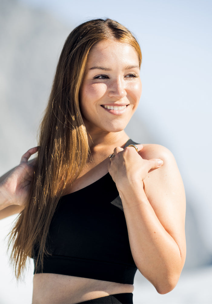 A woman with long, straight brown hair smiles outdoors, wearing the Woolx Livi Bra in Graphite Heather. The moisture-wicking, one-shoulder design stands out as she holds her hair against a bright, softly blurred background.