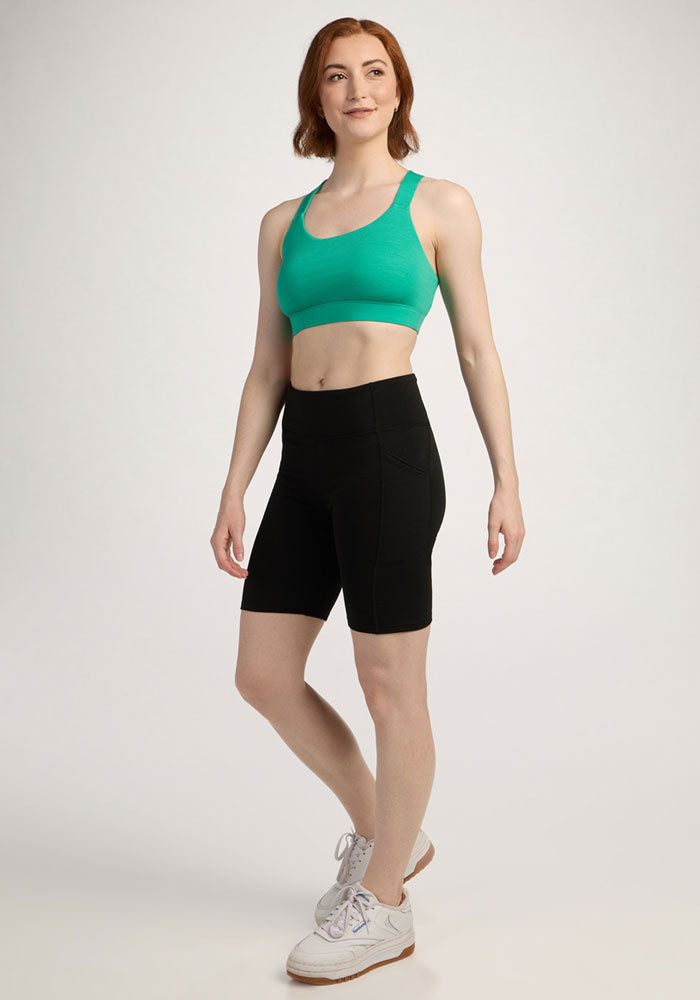 Woman standing and smiling, wearing the Woolx Livi Bra - Jungle with black bike shorts and white sneakers against a plain background.