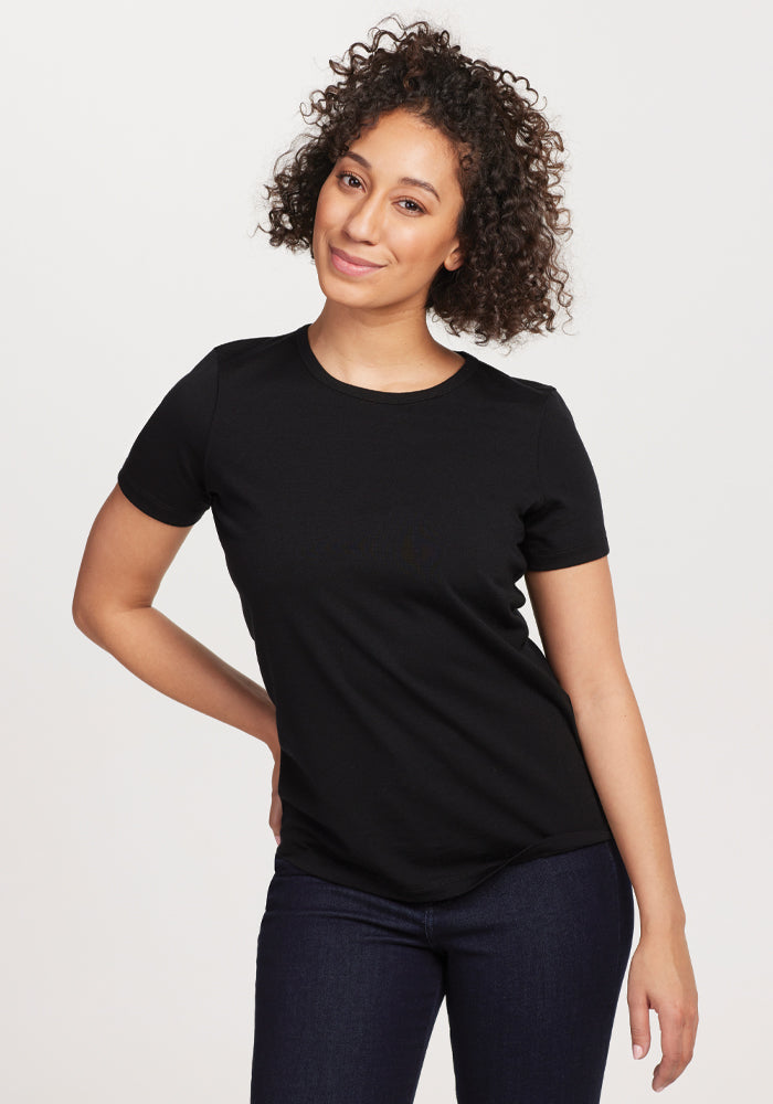 A person with curly hair stands smiling against a plain background, wearing the Woolx Liza Crew Neck Tee in Black made from moisture-wicking fabric, along with dark jeans. One hand rests on their hip while the other hangs by their side. | Tori is 5'7", wearing a size S