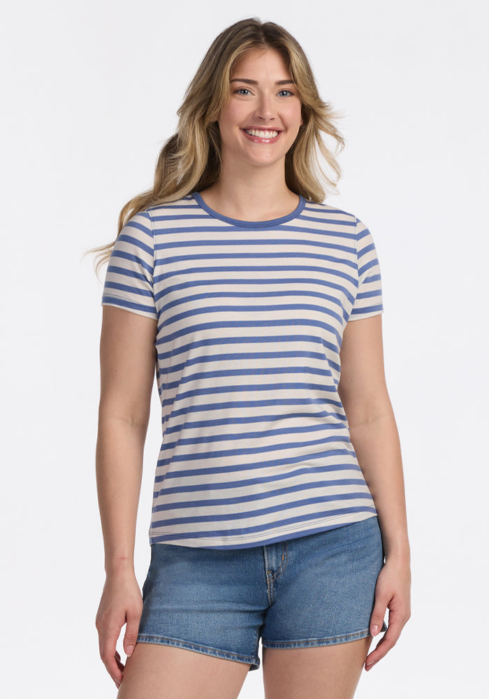 A woman with long, wavy blonde hair smiles in the Woolx Liza Crew Neck Tee - Blue Sand Stripe, paired with denim shorts, standing against a plain white background. | Ariana is 5'11", wearing a size S