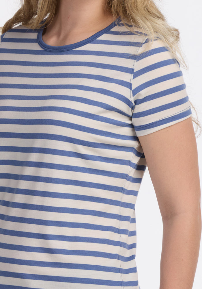 A person with long, wavy blonde hair is shown from the upper torso up, wearing the Woolx Liza Crew Neck Tee in Blue Sand Stripe, a short-sleeved Merino wool shirt with blue and white horizontal stripes.