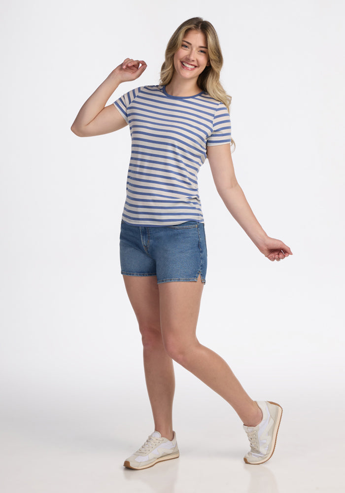 A smiling woman poses in the Woolx Liza Crew Neck Tee - Blue Sand Stripe, paired with denim shorts and white sneakers, against a plain white background.