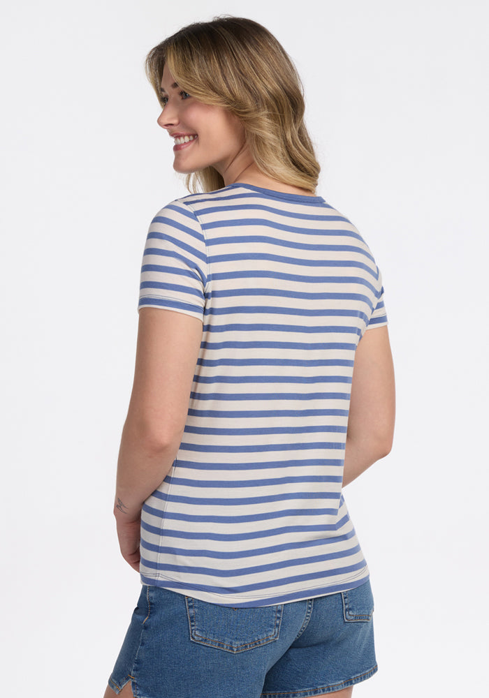 A woman with wavy blonde hair smiles with her back to the camera, wearing the Woolx Liza Crew Neck Tee in Blue Sand Stripe and denim shorts against a plain white background.