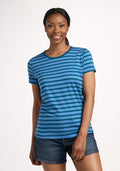 A woman stands against a plain background, smiling. She wears the Woolx Liza Crew Neck Tee - Coastal Stripe and denim shorts. Her hair is pulled back in a low ponytail. | Jade is 5’10”, wearing a size S
