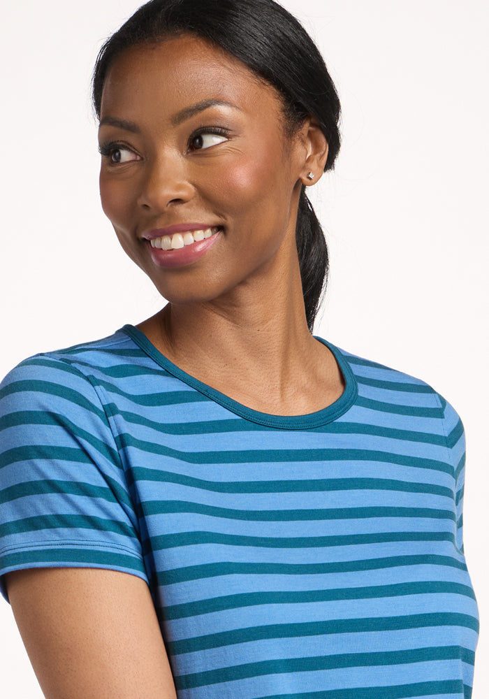 A woman with dark hair in a low ponytail wears the Woolx Liza Crew Neck Tee in Coastal Stripe and smiles while looking slightly to her left, standing against a plain light background.