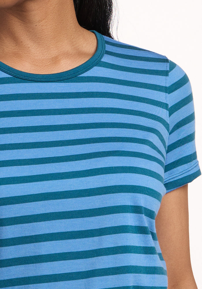 A person wears the Woolx Liza Crew Neck Tee in Coastal Stripe, featuring blue and teal stripes and lightweight performance fabric. Only the lower face, neck, shoulder, and upper chest are visible against a plain light background.