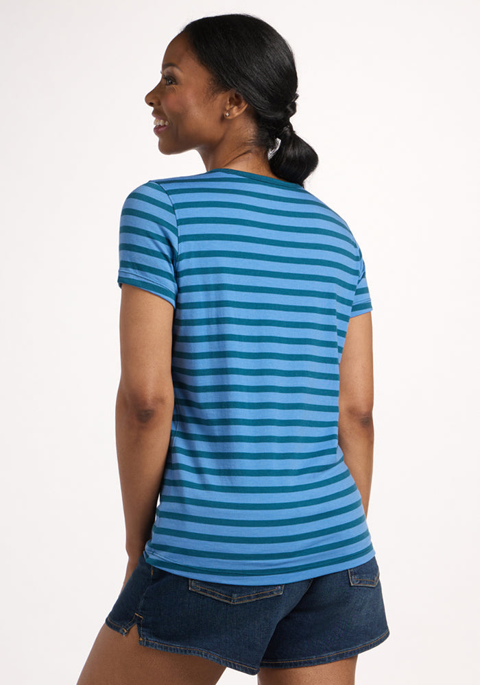 A woman with her hair in a low ponytail stands with her back to the camera, wearing the Woolx Liza Crew Neck Tee in Coastal Stripe and dark denim shorts, against a plain light background.