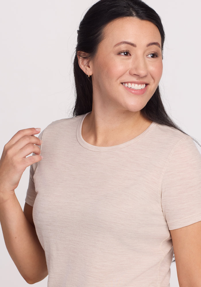 A person with long dark hair wears a Woolx Liza Crew Neck Tee in Cream Heather, smiling while glancing slightly to their right against a plain background.